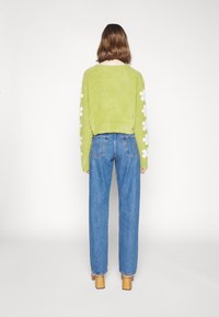 Suéter verde esponjoso con patrones florales blancos en las mangas, combinado con jeans de tiro alto azules y tacones amarillos. Fondo minimalista.
