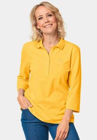 Geel langgevallen poloshirt met kraag, voorzien van een knoopsluiting en geborduurd logo, gecombineerd met blauwe jeans. Zacht katoenen stof.