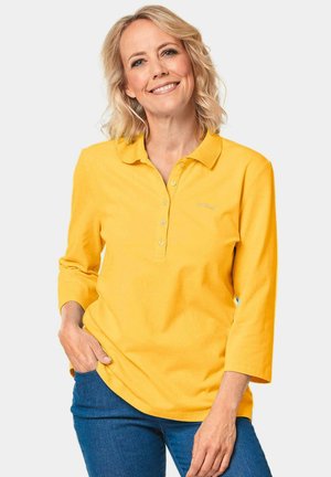 Gelbes langärmliges Poloshirt mit Kragen, ausgestattet mit einer Knopfleiste und besticktem Logo, kombiniert mit blauen Jeans. Aus weichem Baumwollmaterial.