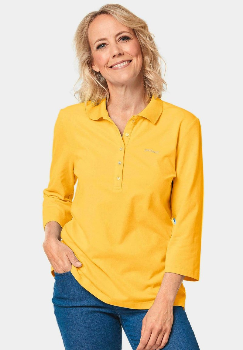 Geel langgevallen poloshirt met kraag, voorzien van een knoopsluiting en geborduurd logo, gecombineerd met blauwe jeans. Zacht katoenen stof.