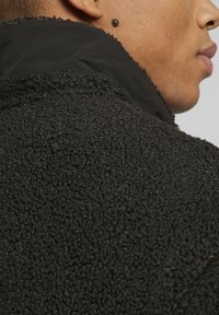 Svart texturerad jacka med en fluffig ytfinish, hög krage och slät innerfoder, som visar upp kontrasterande material och en elegant silhuett.