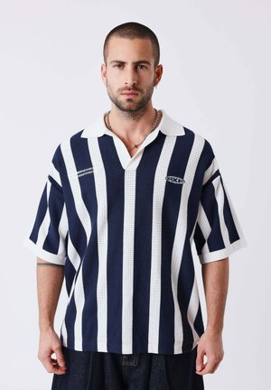 Hombre con cabello corto y barba que lleva una camisa de rugby de rayas verticales en azul marino y blanco con un collar de cadena, de pie frente a un fondo liso.