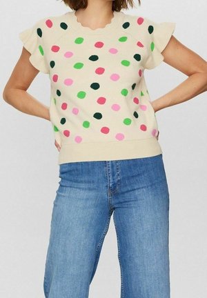 Kvinde iført cremefarvet kortærmet sweater med flerfarvede prikker, kombineret med højtaljede blå jeans, hænderne hvilende på hoften.