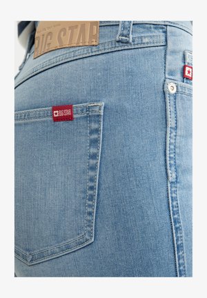 Heledad sinine teksaseelik, mille tagakotis on punane ja valge logo silt, puhta õmbluse ja sileda tekstuuriga.