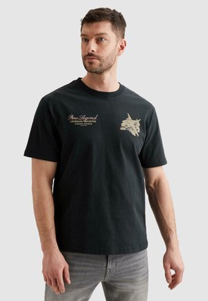 Man wearing black crew neck t-shirt with beige aviation-themed text and graphic on chest, paired with grey jeans, standing against plain background.