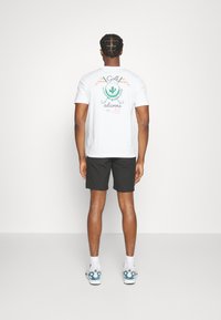 Hvit t-skjorte med et grønt kaktusmotiv og tekst på baksiden, kombinert med svarte shorts og joggesko med blå detaljer.