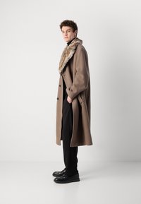Abrigo de mezcla de lana marrón con cuello de piel, de silueta larga y con bolsillos laterales, combinado con pantalones y zapatos negros.