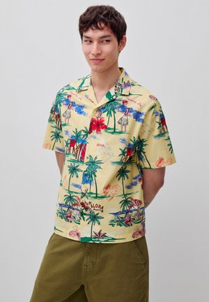 Joven con una camisa hawaiana amarilla de manga corta con palmeras, bailarinas hula, flores rojas de hibisco y pantalones caqui.