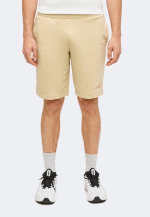 Homme portant un short beige Puma, un t-shirt blanc, des baskets blanches Puma avec des lacets noirs et des chaussettes gris clair, debout face à l’avant.