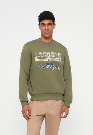 Olivově zelená mikina s vyšitým nápisem "LACOSTE FRENCH SPORTSWEAR" v bílé a šedé barvě, se grafikou hory v modré a bílé.
