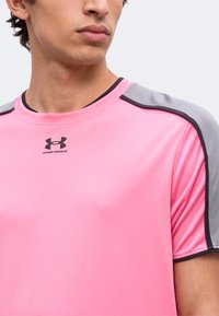 Άνδρας που φοράει ροζ αθλητική μπλούζα Under Armour με γκρι πάνελ στους ώμους και μαύρες λεπτομέρειες, κοιτάζοντας ελαφρώς πλάγια.