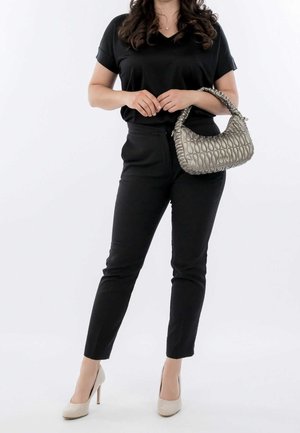 Vrouw in een zwarte outfit en beige hoge hakken die een metalen zilveren gevlochten handtas vasthoudt tegen een witte achtergrond.