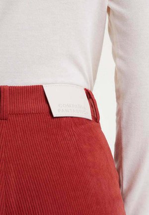 Pantalones de pana rojos con una superficie texturizada y una etiqueta de cuero blanco con una marca en relieve. La parte superior es una camisa de manga larga de tela ligera.
