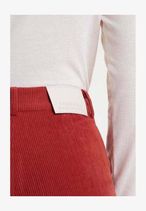 Pantalones de pana rojos con una superficie texturizada y una etiqueta de cuero blanco con una marca en relieve. La parte superior es una camisa de manga larga de tela ligera.