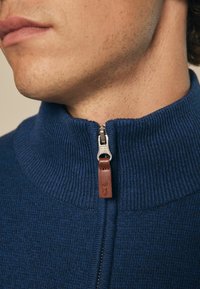 N�ámořnický modrý svetr na zip s žebrovanou texturou, který má stříbrný zip a hnědou koženou úpravu na táhlu zipu.
