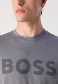 Camiseta de algodón gris con cuello redondo que presenta una gran impresión en gris oscuro de "BOSS" en la parte frontal. Textura suave, diseño ajustado.