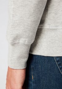 Puño de sudadera gris claro, hecho de tejido texturizado, con un acabado acanalado. Combinado con jeans de mezclilla oscura, mostrando un sutil brillo dorado.
