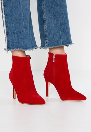 Bottines à talons hauts en daim rouge avec des bouts pointus et des fermetures éclair sur les côtés, associées à un jean bleu en denim avec un ourlet effiloché.