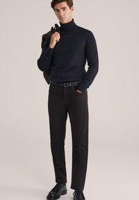 Man in een zwarte turtleneck, donkere broek en zwarte schoenen, met een jas over één schouder, staande tegen een effen achtergrond.