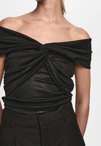 Svart off-shoulder topp med vridet frontdetalj, med en mjuk, glänsande textur. Kombination med högmidjade svarta byxor.