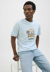 Lichtblauw T-shirt met korte mouwen met een illustratie van een strandhuis, palmbomen en een zonsondergang; katoenen materiaal met een comfortabele pasvorm.
