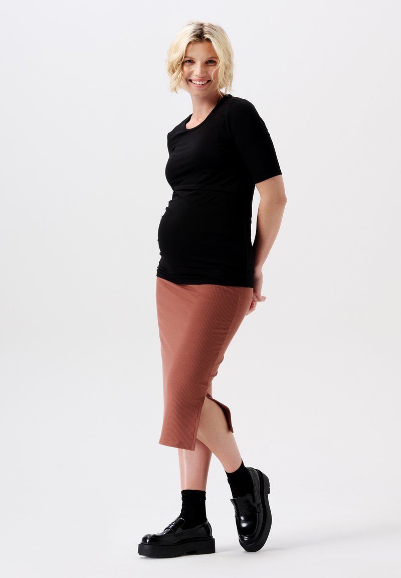 Zwarte, mouwloze zwangerschapsblouse gecombineerd met een geribbelde, middellange, rozenkleurige rok met zijsplitten, aangevuld met zwarte plateau schoenen.