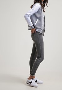 Žena stojící v šedých džínách, bílo-šedé varsity bundě a bílých sneakers Nike s černými logy na pozadí bez detailů.