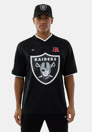 Homme portant un maillot de football noir des Raiders et une casquette assortie des Raiders, debout sur un fond blanc.