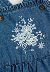 Denim tkanina s volančićima na naramenicama i smešnim dugmadi. Sadrži bijelu cvjetnu vez s detaljnim listovima i naglascima.