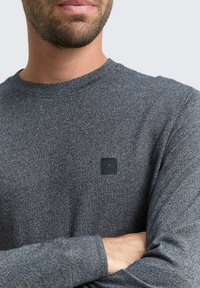 Graues Langarmshirt mit strukturiertem Muster, runder Halsauschnitt und einem schwarzen rechteckigen Logo-Patch auf der linken Brust. Glattes Material.