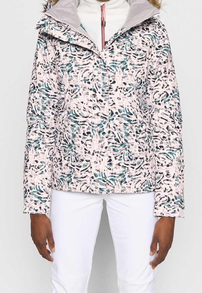 Femme portant une veste à zip à motifs avec des taches turquoise, noires et blanches sur fond blanc, associée à un pantalon blanc.