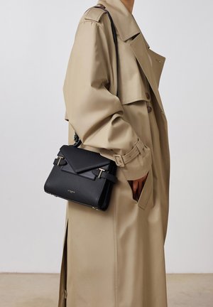 Sac à main en cuir texturé noir avec fermeture à rabat angulaire, accents en matériel doré et bandoulière amovible, tenu contre un trench-coat beige clair.