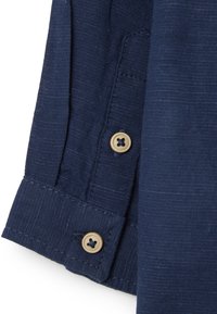 Detalle de camisa de manga larga azul marino, con tejido texturizado, puño abotonado con botón beige y costura en contraste.
