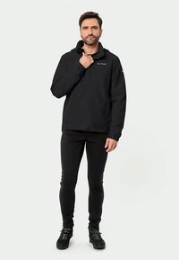 Vyras, dėvintis juodą lauko striukę, juodas kelnes ir juodus žygių batus, stovi prieš vienodą šviesią fono.