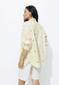 Chemise boutonnière jaune clair avec petite broderie florale, coupe décontractée, manches longues et ourlet arrondi, assortie à un short blanc.