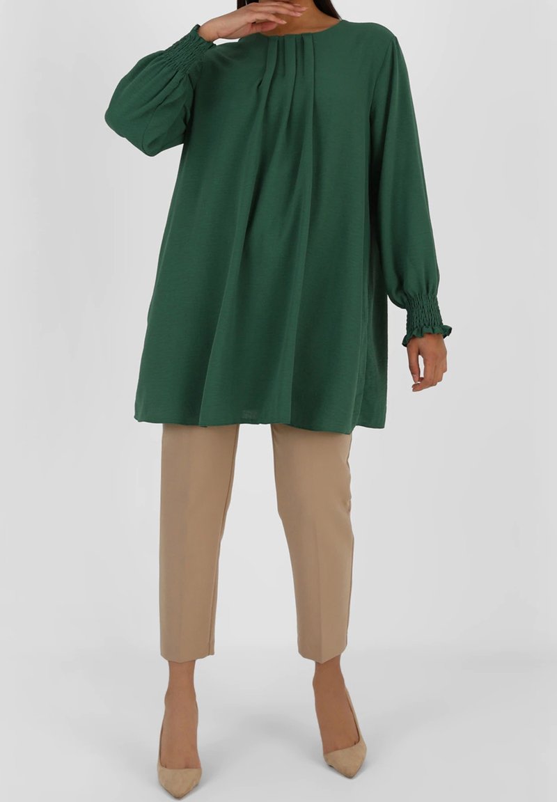 Femme portant une tunique verte ample à manches longues avec poignets froncés, pantalon beige raccourci et escarpins beige à bouts pointus.