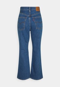 Jean en denim bleu taille haute avec poches arrière, jambes évasées, coutures marron et écusson en cuir de la marque sur la ceinture.