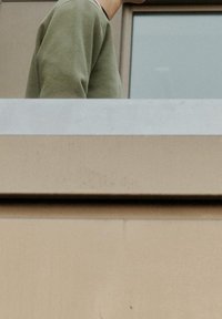 Felpa verde oliva con scollo rotondo, indossata da una persona in piedi vicino a una finestra, caratterizzata da un design minimalista e una texture morbida.