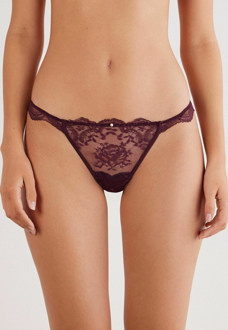 String en dentelle bordeaux avec des motifs floraux, des bords festonnés et une petite gemme décorative à l'avant. Matière douce et légère.