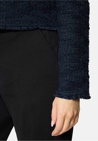 Main d'une femme près de la poche latérale d'un pantalon noir, portant un haut à manches longues bleu foncé texturé avec des poignets effilochés.