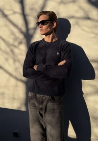 Junger Mann mit schwarzen Sonnenbrillen, marineblauem Pullover und grauen Jeans steht mit verschränkten Armen vor einer sonnenbeschienenen Wand, die Schatten von Baumzweigen wirft.