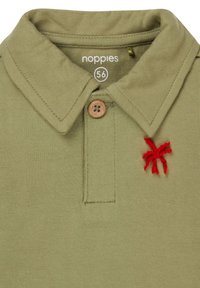 Oliwkowa koszulka polo z kołnierzykiem, zamykana na drewniany guzik, z czerwonym haftem w kształcie pająka po lewej stronie klatki piersiowej. Widoczna metka rozmiaru 56.