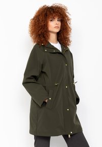 Giacca impermeabile verde oliva con cappuccio, chiusura a pressione e zip, vita regolabile con coulisse e due tasche laterali. Accenti dorati.