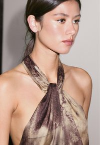 Hnědý a béžový šátek se vzorem a uzlovým designem kolem krku, s hedvábnou texturou a splývavým drapériem.