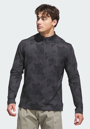 Nuori mies, joka pukeutunut mustaan camokuvioiseen pitkähiharaiseen poolopuseroon, jossa on neljänneksen vetoketju. Hän seisoo rennosti käsivarret alas, katse vasemmalle.