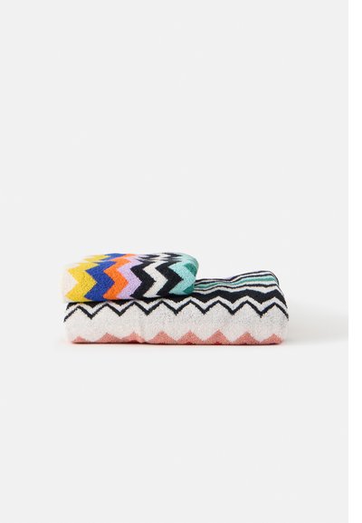 Deux serviettes pliées empilées, toutes deux avec des motifs en zigzag ; la serviette du dessus a des zigzags multicolores, celle du dessous des zigzags noirs et roses.
