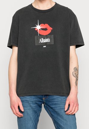 Person iført en sort AllSaints T-shirt med et rødt læbe-print, en hvid stjerne og teksten "Tokyo" og "2022," parret med blå jeans og en slange-tatovering.