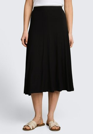 Frau, die einen schwarzen Midi-Rock, ein weißes Top und beige Sandalen trägt und vor einem einfarbigen hellen Hintergrund steht.