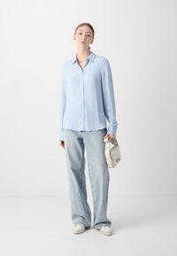 Camicia azzurra chiara a maniche lunghe con righe verticali, abbinata a jeans chiari a taglio largo e sneakers bianche. Tiene in mano una borsa color crema.