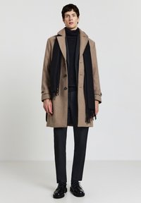 Cappotto di lana marrone con un ampio colletto, dolcevita nero e sciarpa nera con frange, abbinati a pantaloni scuri attillati e scarpe nere.